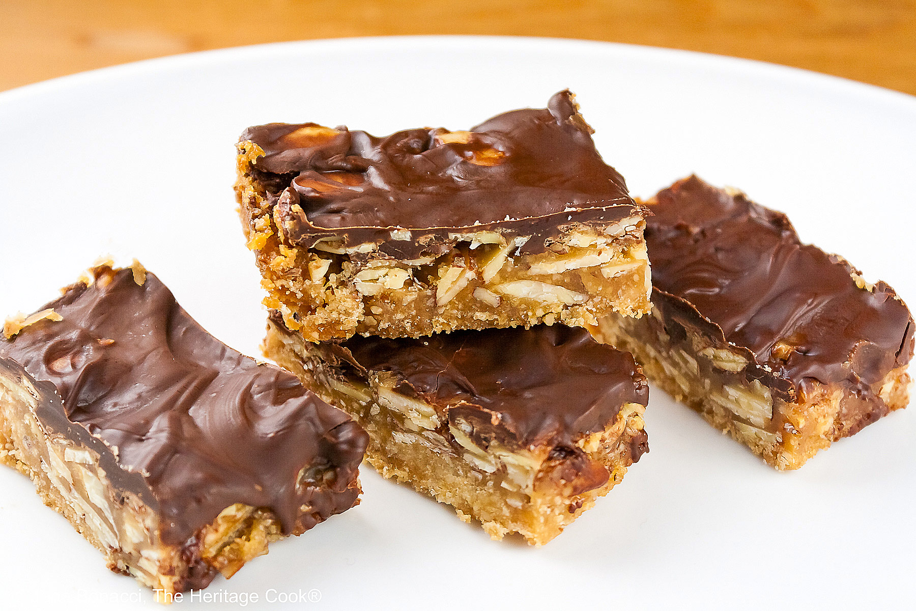 Luscious Brown Sugar Almond Bars, layered with a bottom crust, caramel filling, and topped with melted chocolate, stacked or scattered on a white plate, sitting on a golden wooden table © 2025 Jane Bonacci, The Heritage Cook.