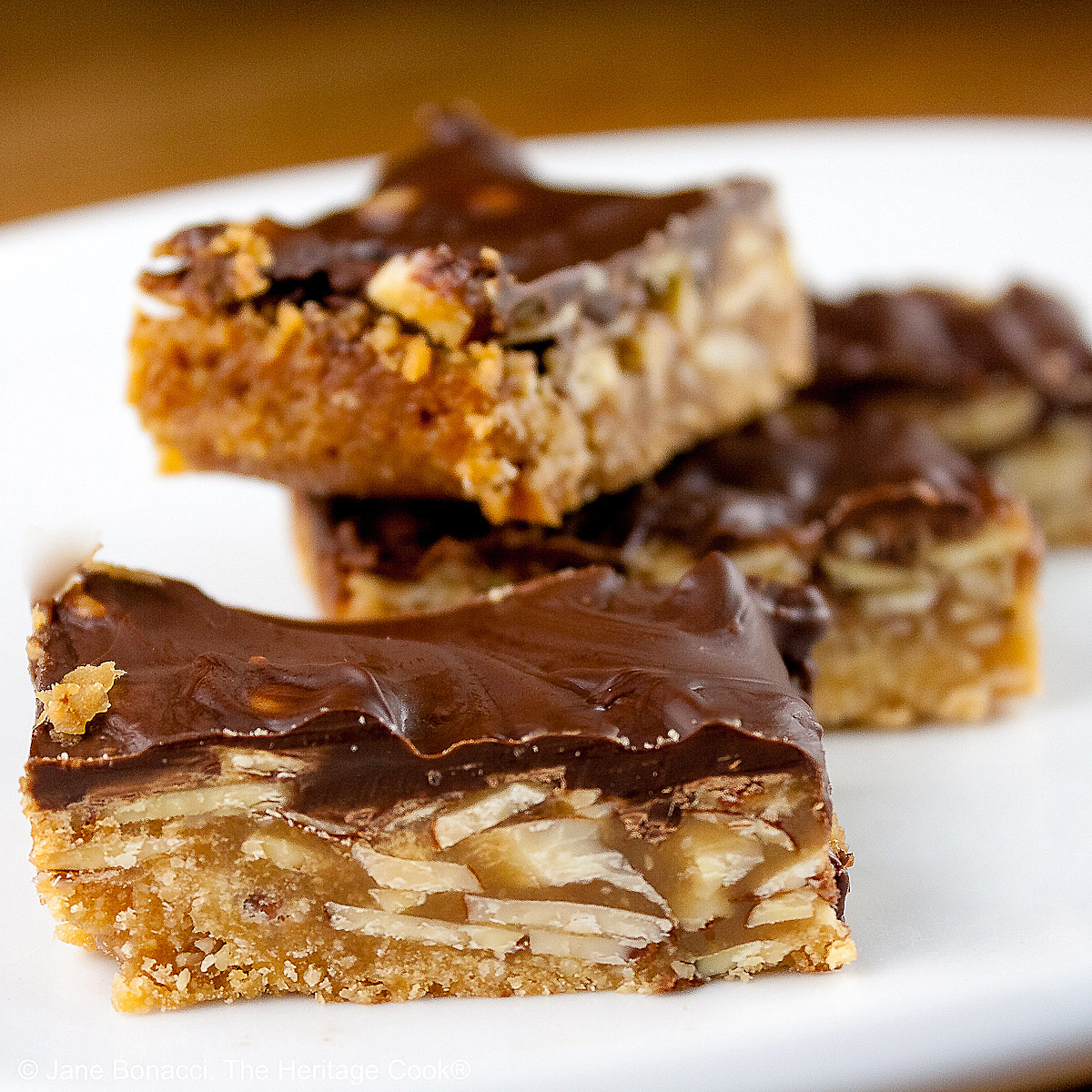 Luscious Brown Sugar Almond Bars, layered with a bottom crust, caramel filling, and topped with melted chocolate, stacked or scattered on a white plate, sitting on a golden wooden table © 2025 Jane Bonacci, The Heritage Cook.