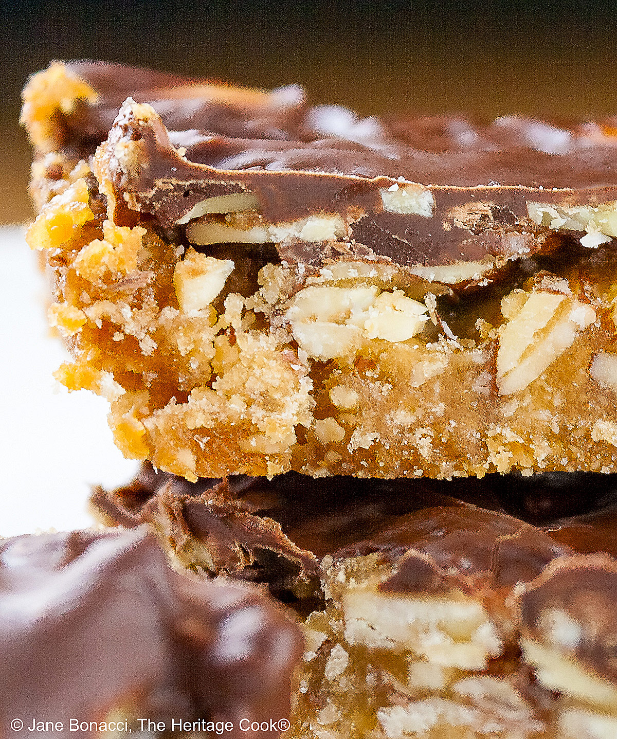 Luscious Brown Sugar Almond Bars, layered with a bottom crust, caramel filling, and topped with melted chocolate, stacked or scattered on a white plate, sitting on a golden wooden table © 2025 Jane Bonacci, The Heritage Cook.