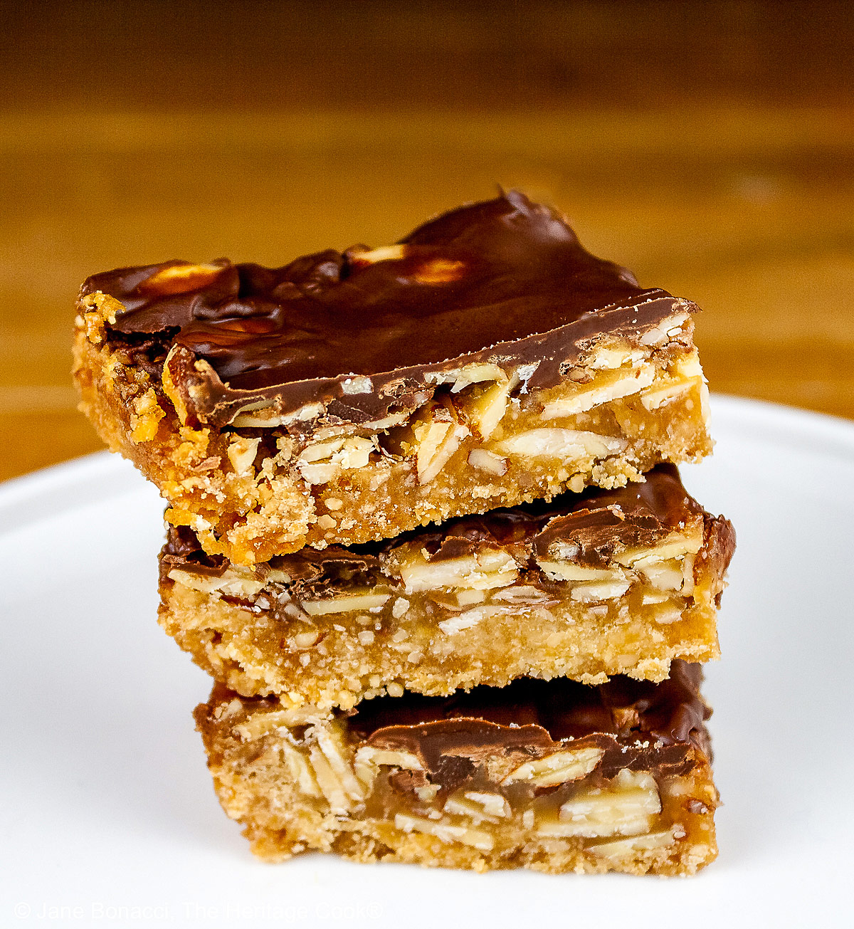 Luscious Brown Sugar Almond Bars, layered with a bottom crust, caramel filling, and topped with melted chocolate, stacked or scattered on a white plate, sitting on a golden wooden table © 2025 Jane Bonacci, The Heritage Cook.