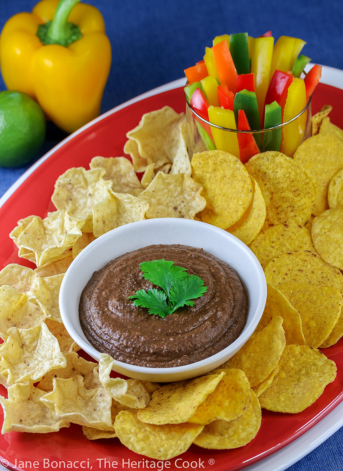 Bowl of black bean dip topped with cilantro leaves and surrounded by tortilla chips and bell pepper strips for dipping; Chipotle Black Bean Dip for Super Bowl Sunday © 2023 Jane Bonacci, The Heritage Cook.