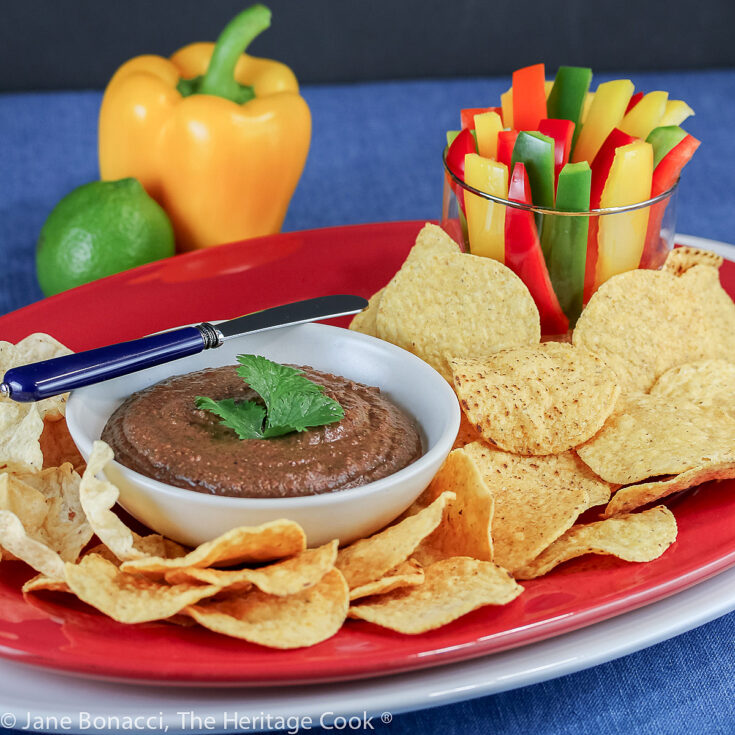 Bowl of black bean dip topped with cilantro leaves and surrounded by tortilla chips and bell pepper strips for dipping; Chipotle Black Bean Dip for Super Bowl Sunday © 2023 Jane Bonacci, The Heritage Cook.