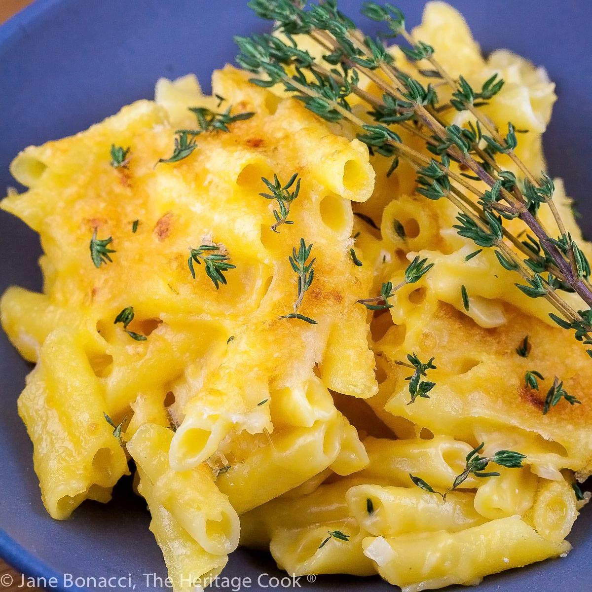 Yellow mac and cheese on a blue plate with green thyme sprigs on top; Cheese Fondue Mac and Cheese © 2022 Jane Bonacci, The Heritage Cook. 