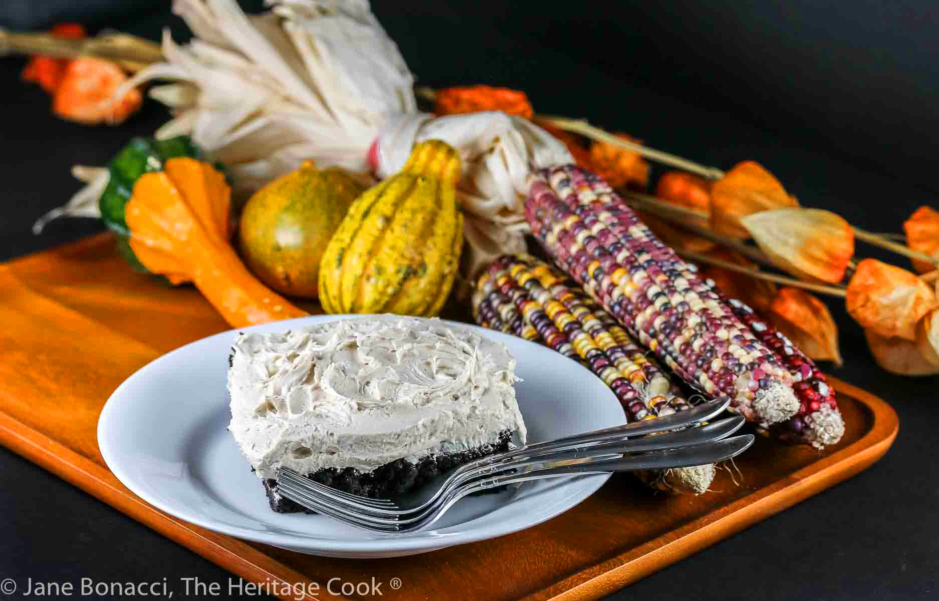 Square of chocolate cake topped with pillows of white maple frosting on a plate in front of seasonal corn cobs, flowers, gourds, etc. © 2023 Jane Bonacci, The Heritage Cook.
