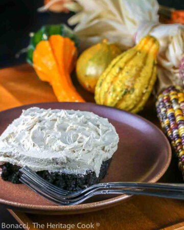 Square of chocolate cake topped with pillows of white maple frosting on a plate in front of seasonal corn cobs, flowers, gourds, etc. © 2023 Jane Bonacci, The Heritage Cook.