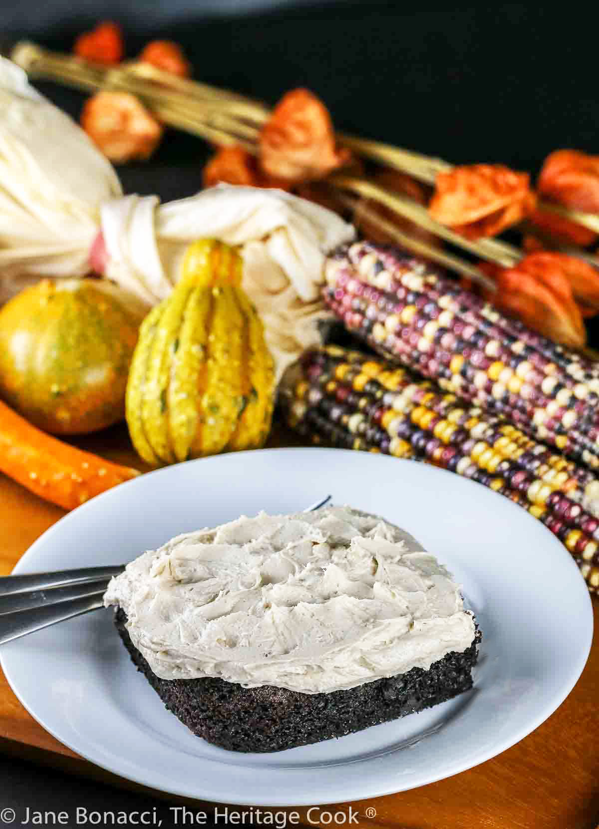 Square of chocolate cake topped with pillows of white maple frosting on a plate in front of seasonal corn cobs, flowers, gourds, etc. © 2023 Jane Bonacci, The Heritage Cook.