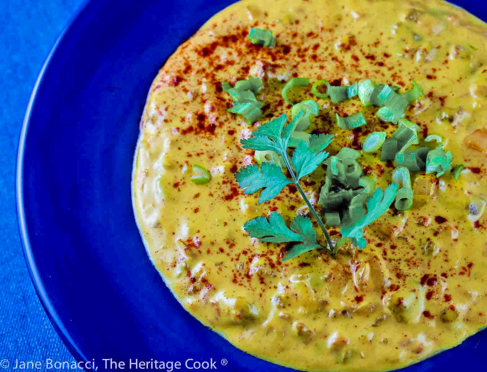 Deep blue bowl filled with yellow cheese dip with chunks of chorizo in it and cilantro on top, sitting on a denim cloth © 2024 Jane Bonacci, The Heritage Cook. 