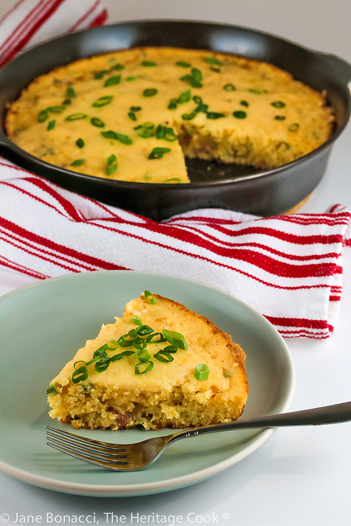 Ham and Cheese Cornbread baked in a cast iron skillet, slices on a small plate topped with sliced green onions © 2023 Jane Bonacci, The Heritage Cook.