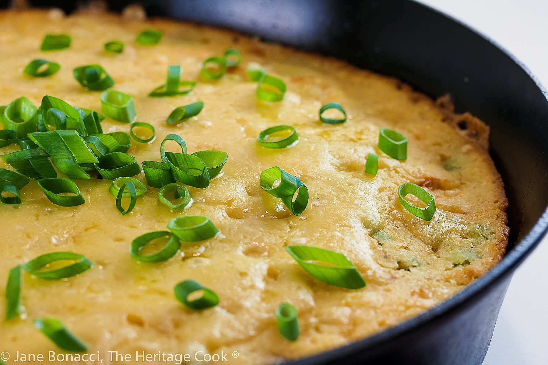Close up of Ham and Cheese Cornbread baked in a cast iron skillet, slices on a small plate topped with sliced green onions © 2023 Jane Bonacci, The Heritage Cook.