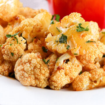 Large pile of orange colored (from the sauce) Grilled Buffalo Cauliflower on a white platter with a red bowl of blue cheese dipping sauce, everything sprinkled with chopped parsley © 2024 Jane Bonacci, The Heritage Cook.