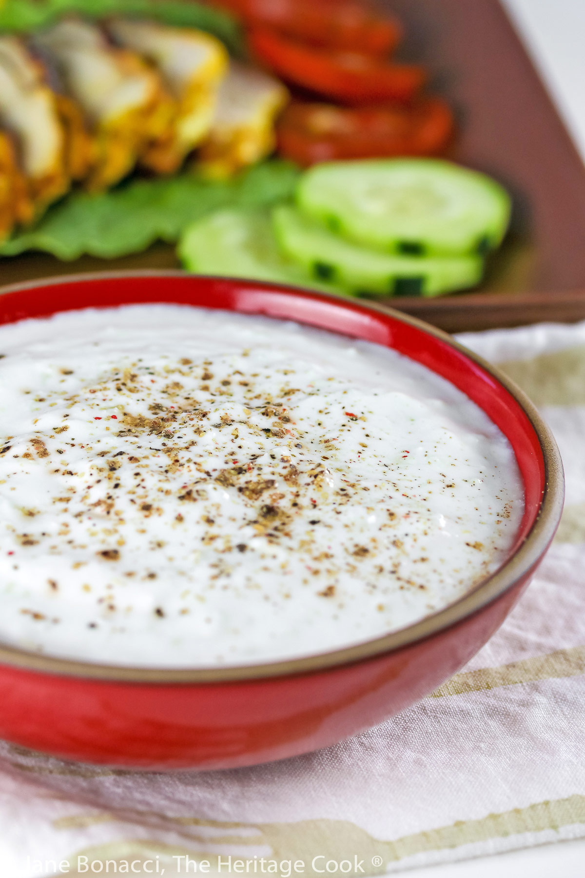 Close up of cucumber-yogurt sauce sprinkled with ground pepper in red bowl. 