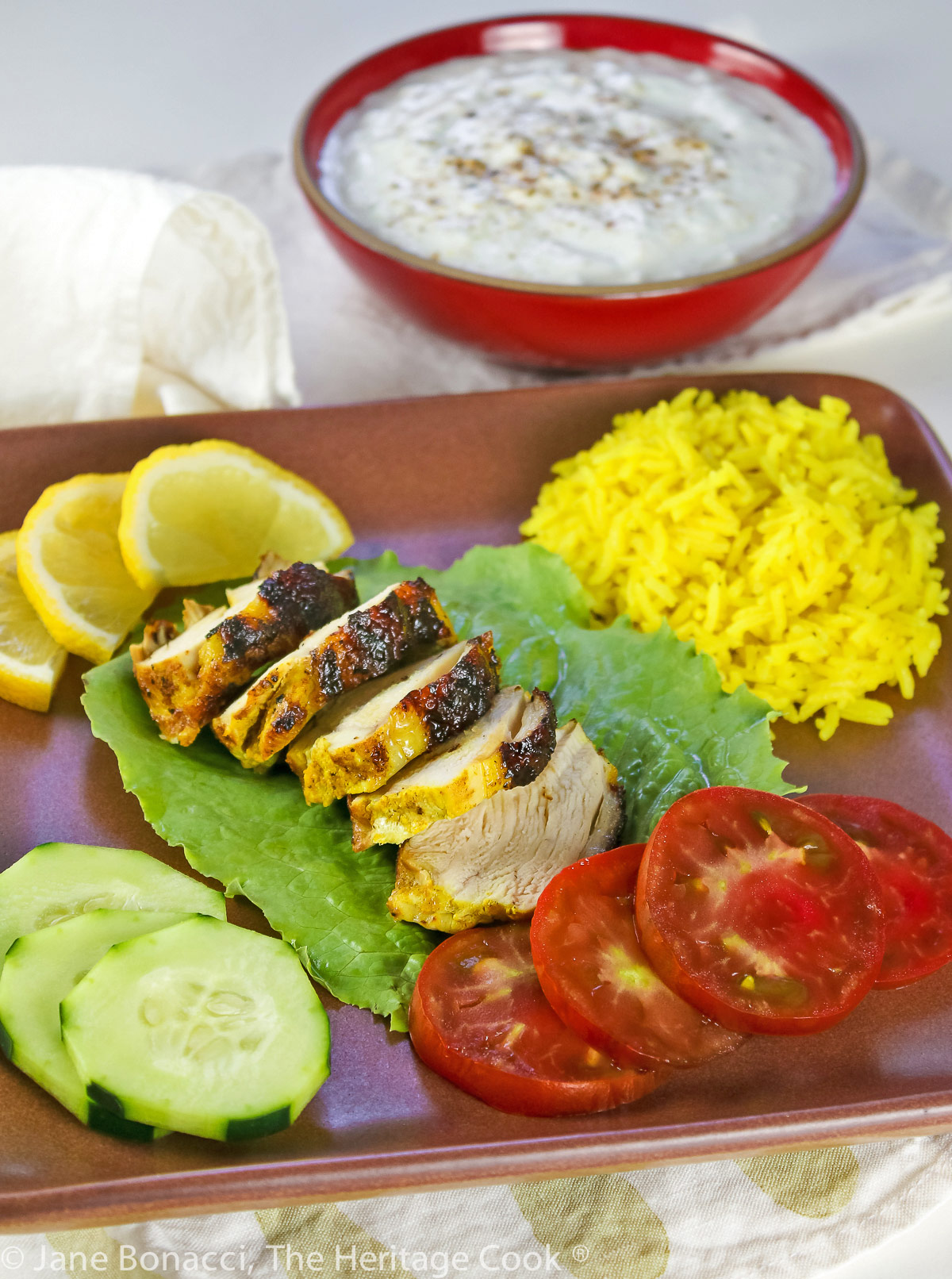 Grilled chicken breast sliced thickly and served on a bed of lettuce with yellow rice, vegetables, and a bowl of cooling yogurt sauce; Tandoori Grilled Chicken with Cucumber Yogurt Sauce (Gluten-Free) © 2023 Jane Bonacci, The Heritage Cook.