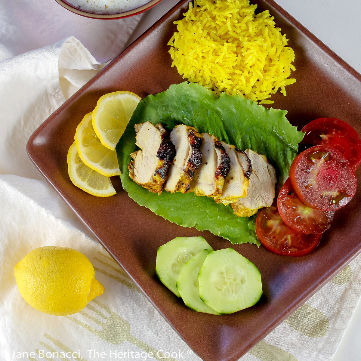 Grilled chicken breast sliced thickly and served on a bed of lettuce with yellow rice, vegetables, and a bowl of cooling yogurt sauce; Tandoori Grilled Chicken with Cucumber Yogurt Sauce (Gluten-Free) © 2023 Jane Bonacci, The Heritage Cook.