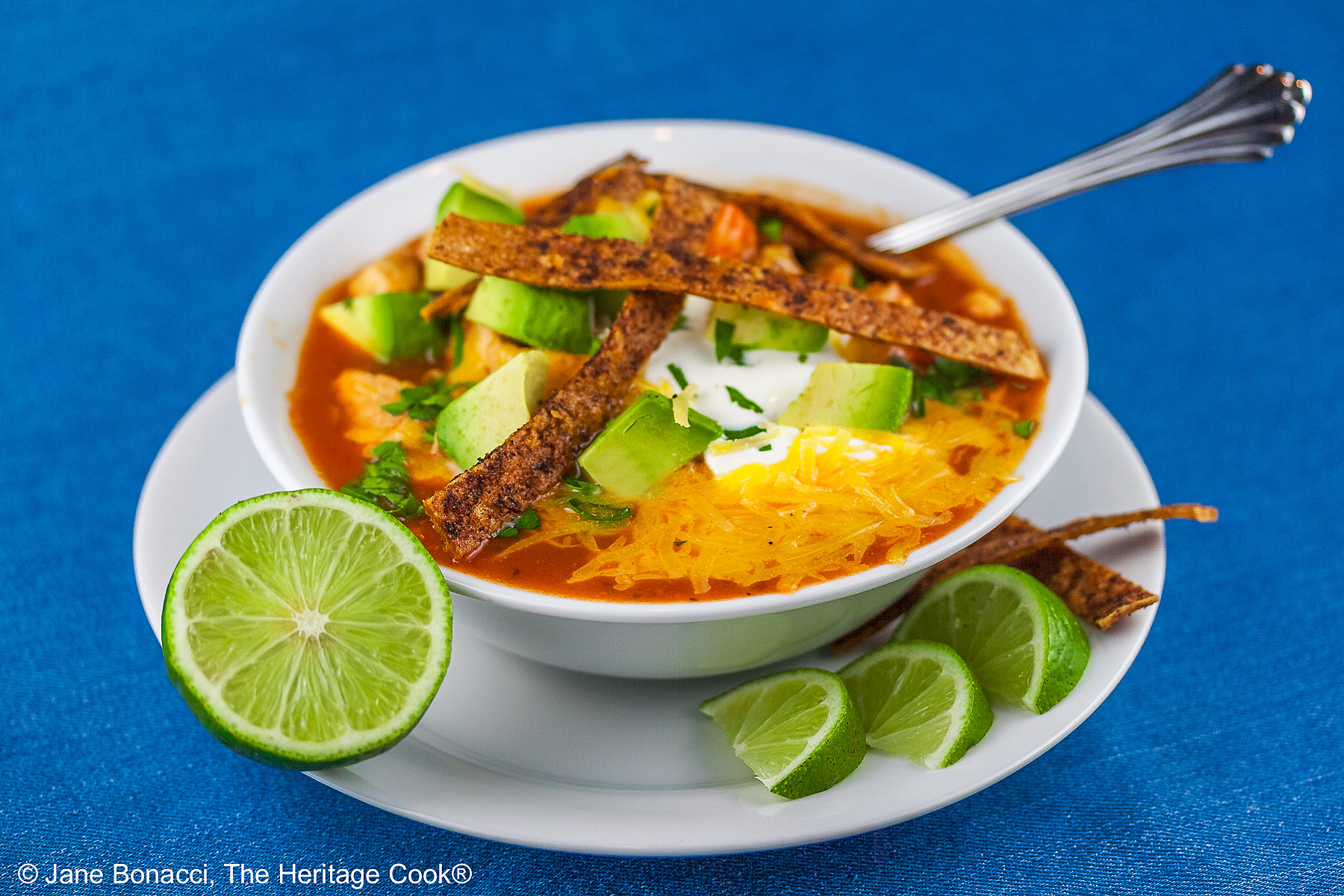 White bowl sitting on a white plate on a blue background, filled with rich, deep red chicken chile soup with spiced tortilla strips, sour cream, avocado cubes, shredded cheese, chopped cilantro, green onions - beautiful toppings add luscious flavor and texture, with lime wedges alongside © 2025 Jane Bonacci, The Heritage Cook.