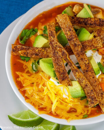 White bowl sitting on a white plate on a blue background, filled with rich, deep red chicken chile soup with spiced tortilla strips, sour cream, avocado cubes, shredded cheese, chopped cilantro, green onions - beautiful toppings add luscious flavor and texture, with lime wedges alongside © 2025 Jane Bonacci, The Heritage Cook.