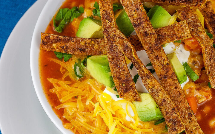 White bowl sitting on a white plate on a blue background, filled with rich, deep red chicken chile soup with spiced tortilla strips, sour cream, avocado cubes, shredded cheese, chopped cilantro, green onions - beautiful toppings add luscious flavor and texture, with lime wedges alongside © 2025 Jane Bonacci, The Heritage Cook.