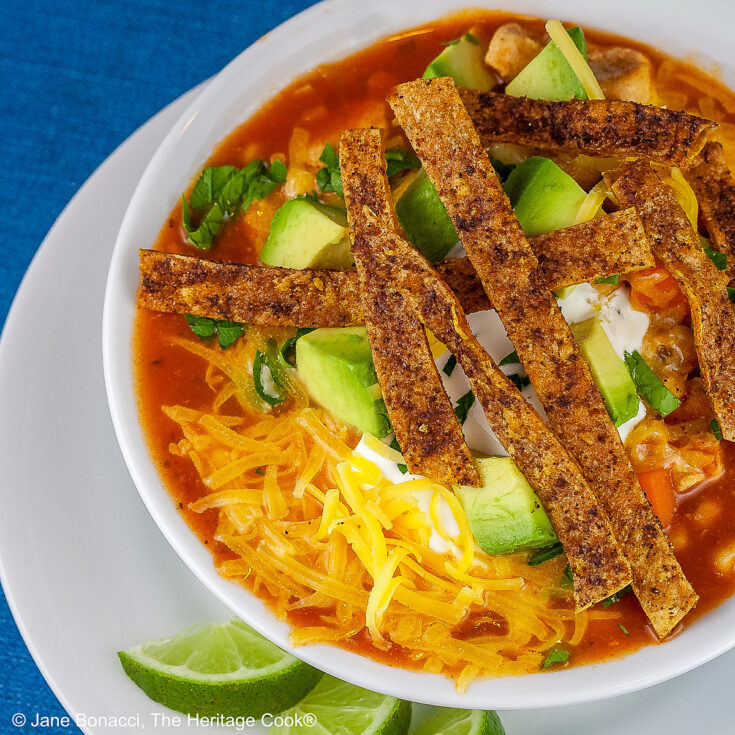 White bowl sitting on a white plate on a blue background, filled with rich, deep red chicken chile soup with spiced tortilla strips, sour cream, avocado cubes, shredded cheese, chopped cilantro, green onions - beautiful toppings add luscious flavor and texture, with lime wedges alongside © 2025 Jane Bonacci, The Heritage Cook.