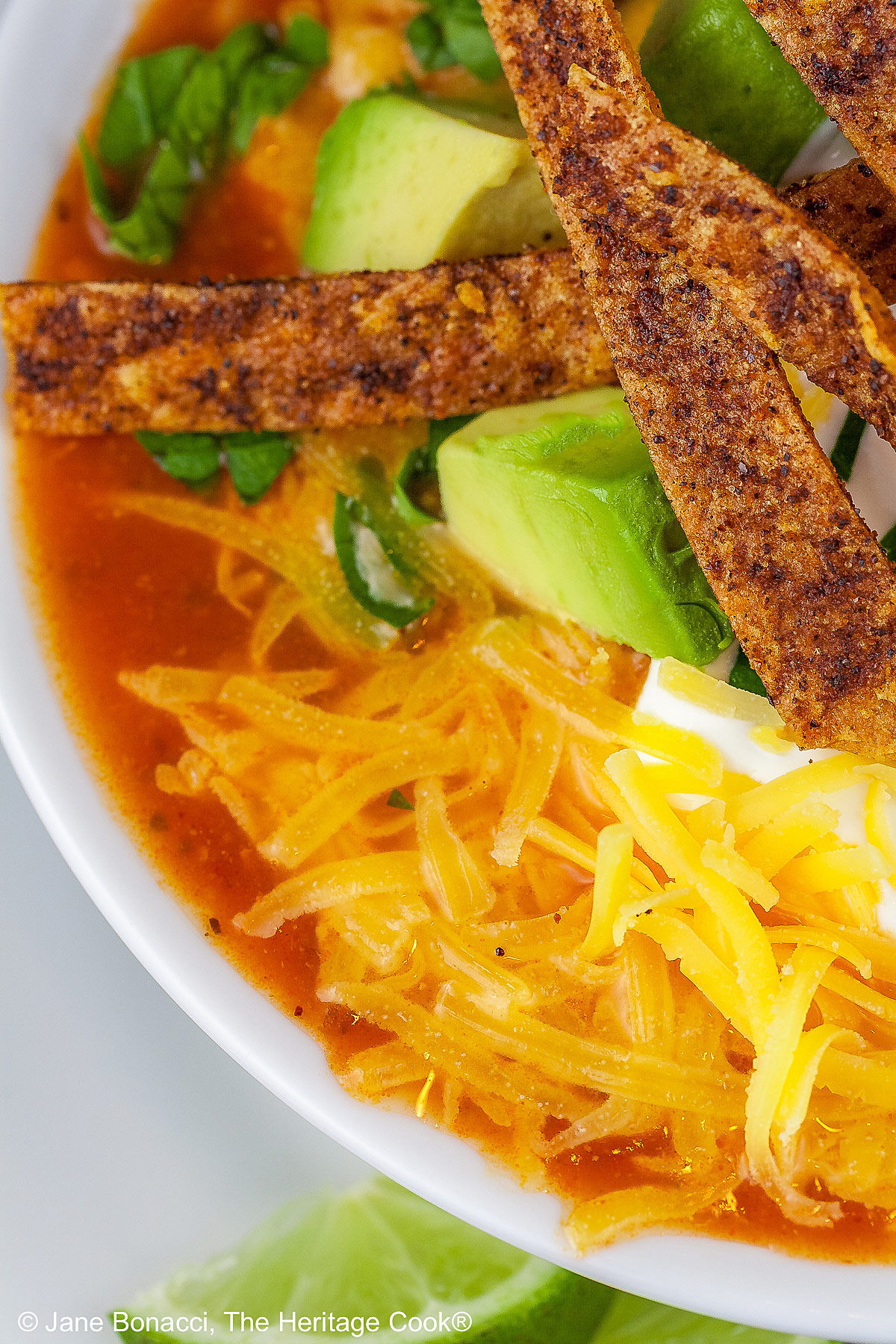 White bowl sitting on a white plate on a blue background, filled with rich, deep red chicken chile soup with spiced tortilla strips, sour cream, avocado cubes, shredded cheese, chopped cilantro, green onions - beautiful toppings add luscious flavor and texture, with lime wedges alongside © 2025 Jane Bonacci, The Heritage Cook.