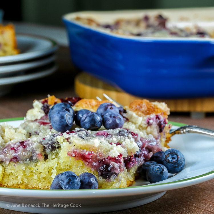 slice of blueberry buckle with baking pan in the back; Fresh Blueberry Buckle from the America the Great Cookbook © 2018 Jane Bonacci, The Heritage Cook