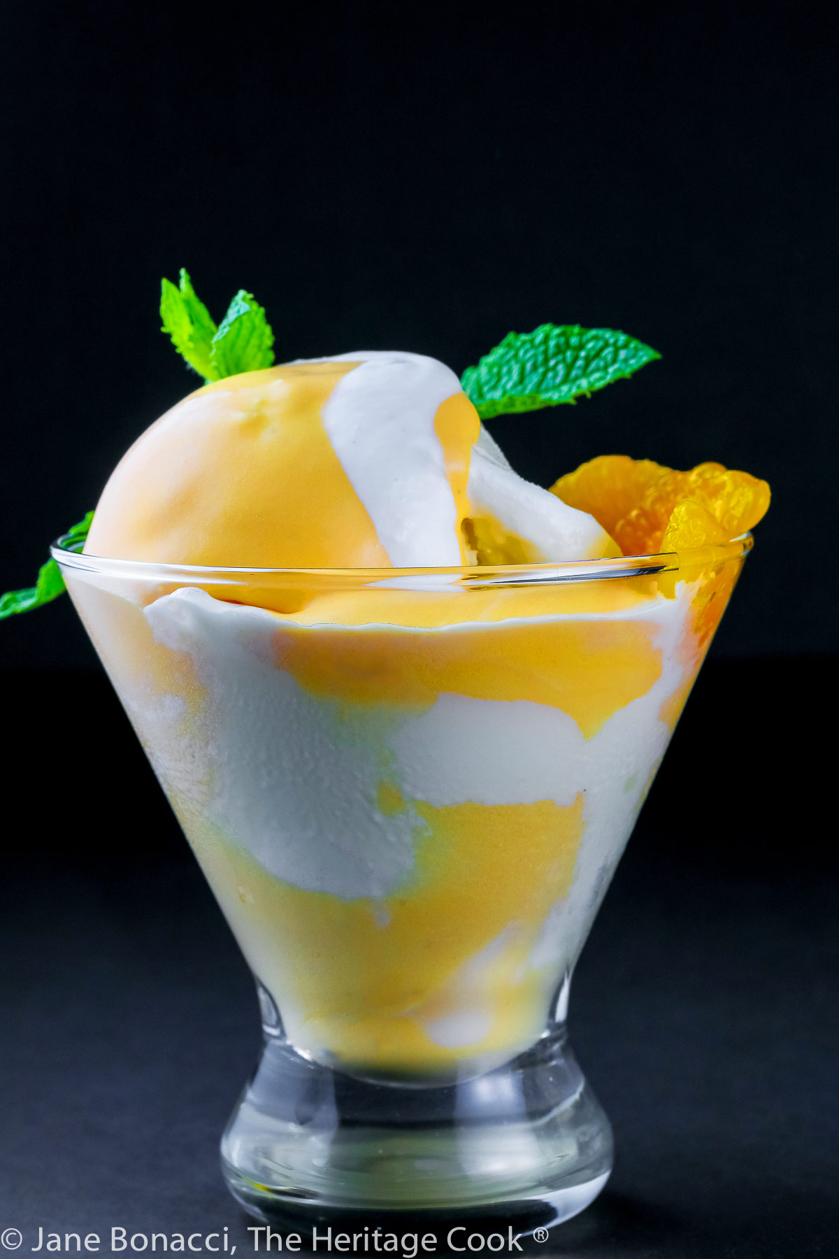 Vanilla ice cream with orange Creamsicle mousse in glass dish topped with whipped cream and a sprig of mint; on black background © 2023 Jane Bonacci, The Heritage Cook. 