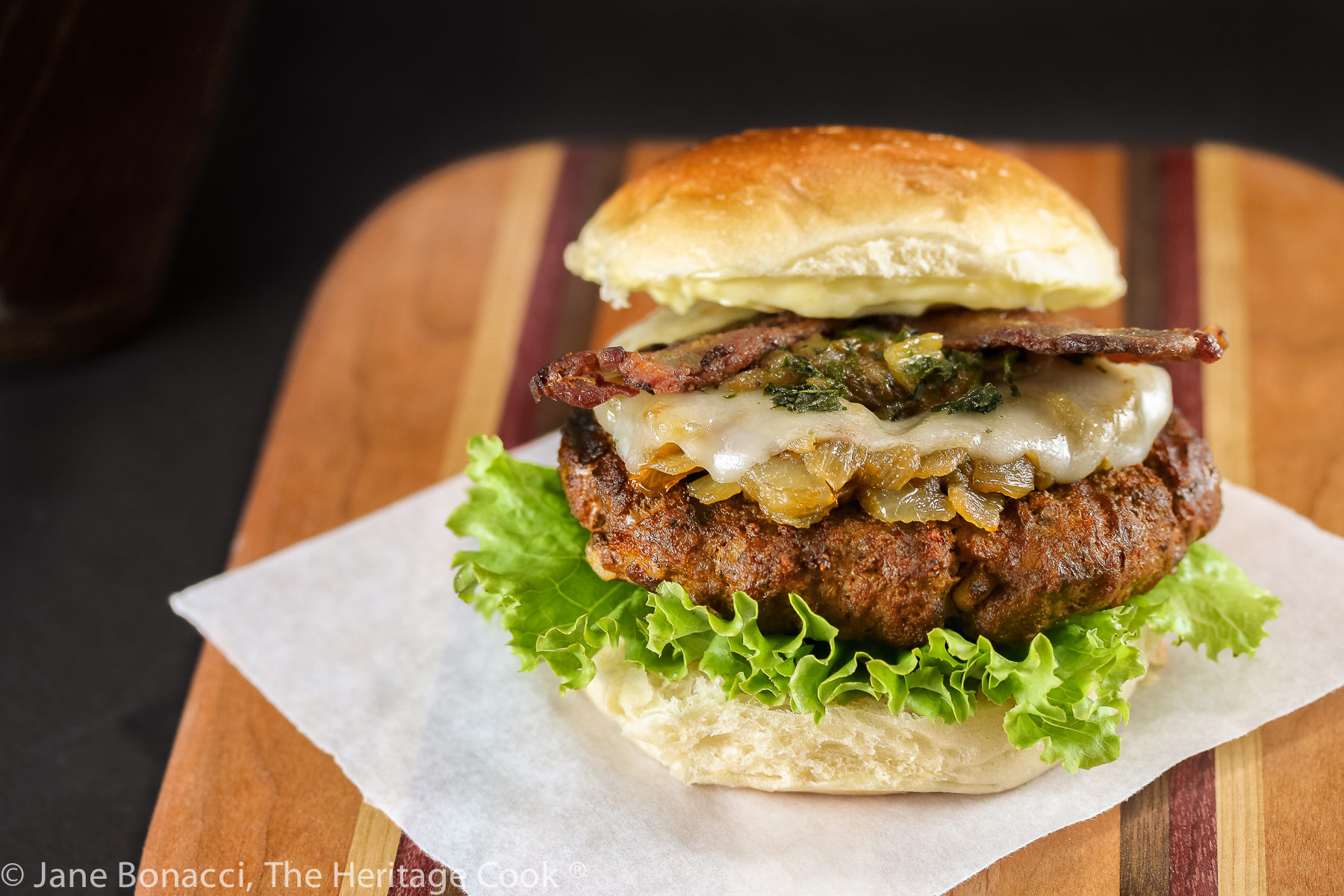 A grilled buffalo burger studded with chopped bacon fat and layered with lettuce, bacon, melted cheese, sauteed onions and Hatch green chiles; BBQ Green Chile Bacon Burgers © 2023 Jane Bonacci, The Heritage Cook. 