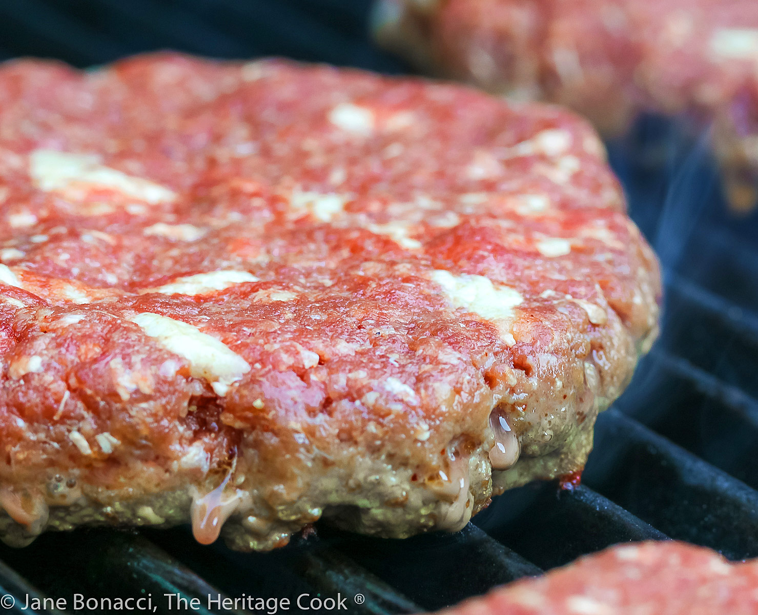Hamburger patty cooking on the grill, showing the pieces of bacon fat. 