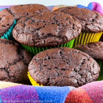 Chocolate muffins with high rounded tops in multi-colored muffin papers and stacked in a basket lined with a brightly colored napkin © 2023 Jane Bonacci, The Heritage Cook.