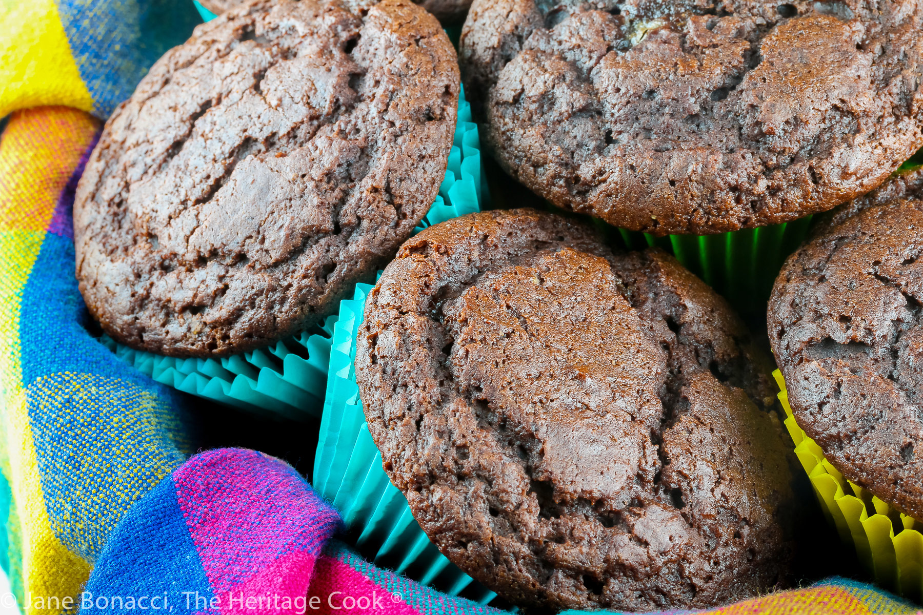 Chocolate muffins with high rounded tops in multi-colored muffin papers and stacked in a basket lined with a brightly colored napkin © 2023 Jane Bonacci, The Heritage Cook. 