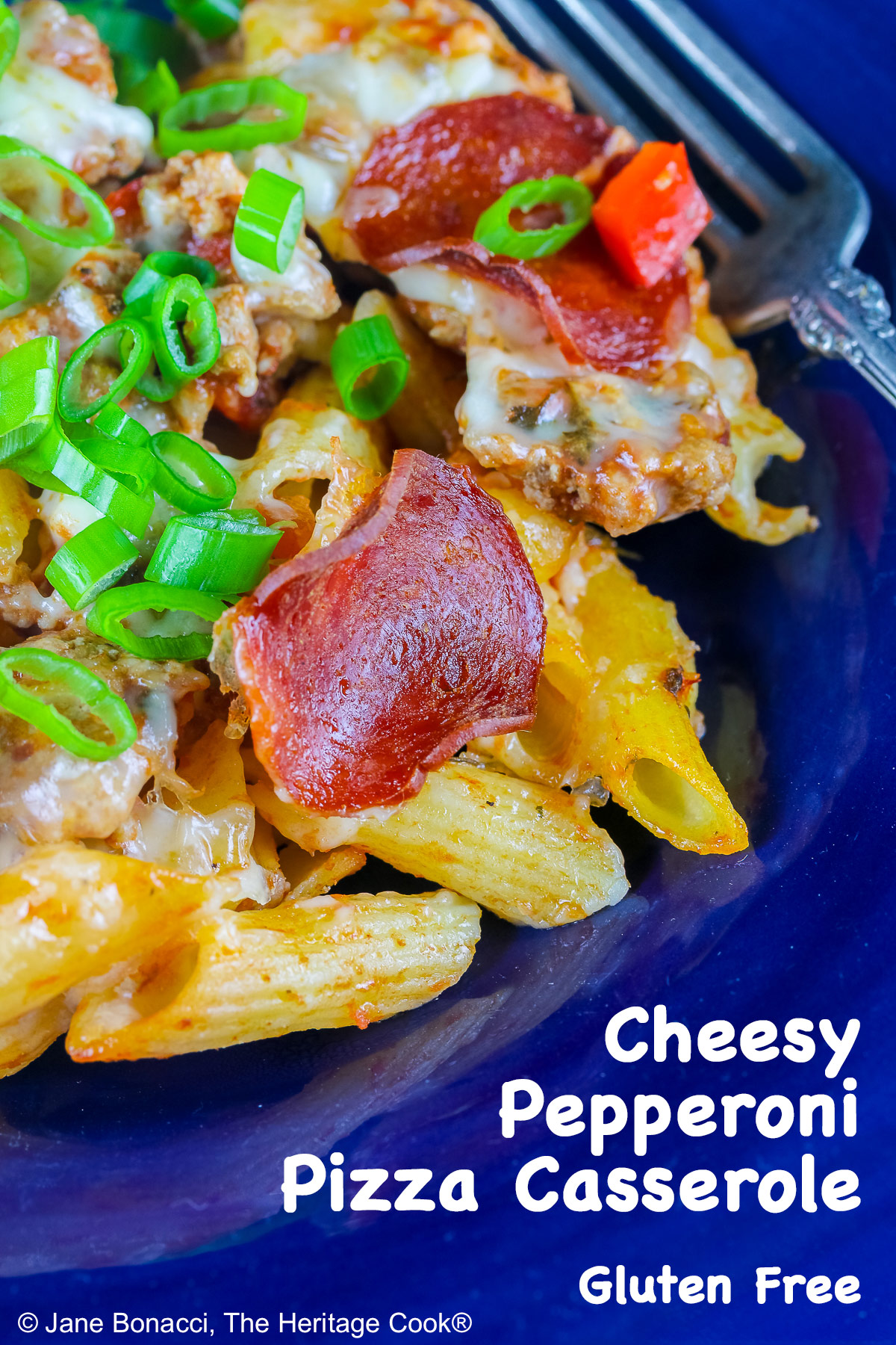 Cheesy Pepperoni Pizza Casserole scooped into a dark blue bowl with a wide rim, topped with pepperoni slices and sliced green onions, sitting on a blue and white cloth © 2026 Jane Bonacci, The Heritage Cook.