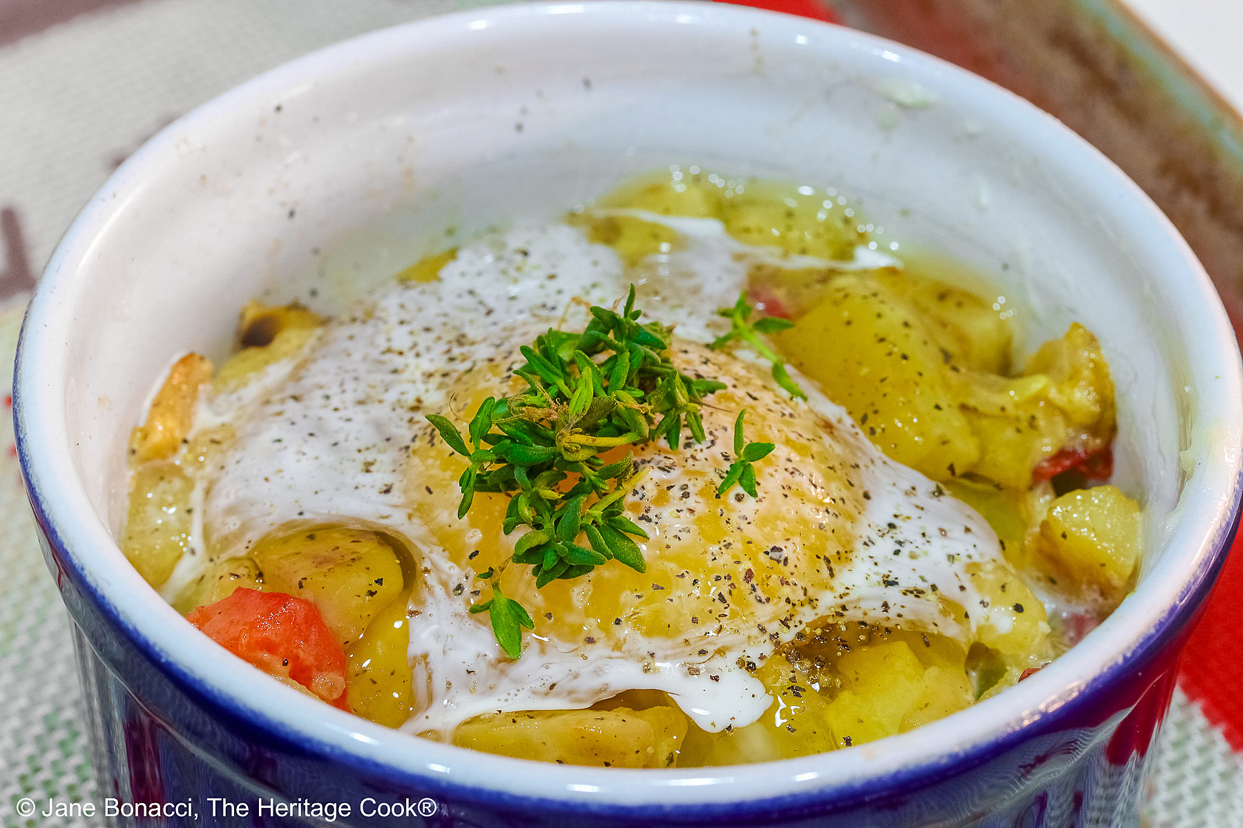 Provençal baked eggs in a potato nest in dark blue ramekins on a baking sheet with a Silpat to stop sliding, sitting on a multi-colored cloth with a black spoon © 2026 Jane Bonacci, The Heritage Cook.
