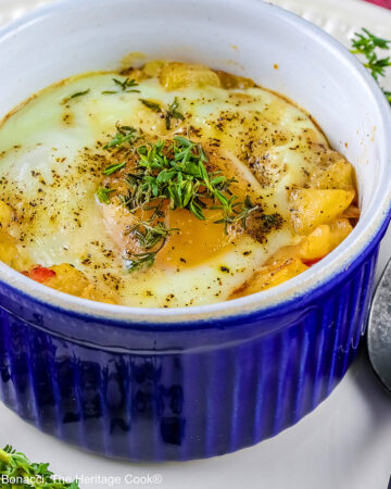 Provençal baked eggs in a potato nest in dark blue ramekins on a baking sheet with a Silpat to stop sliding, sitting on a multi-colored cloth with a black spoon © 2026 Jane Bonacci, The Heritage Cook.