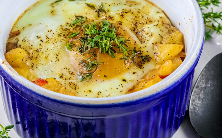 Provençal baked eggs in a potato nest in dark blue ramekins on a baking sheet with a Silpat to stop sliding, sitting on a multi-colored cloth with a black spoon © 2026 Jane Bonacci, The Heritage Cook.