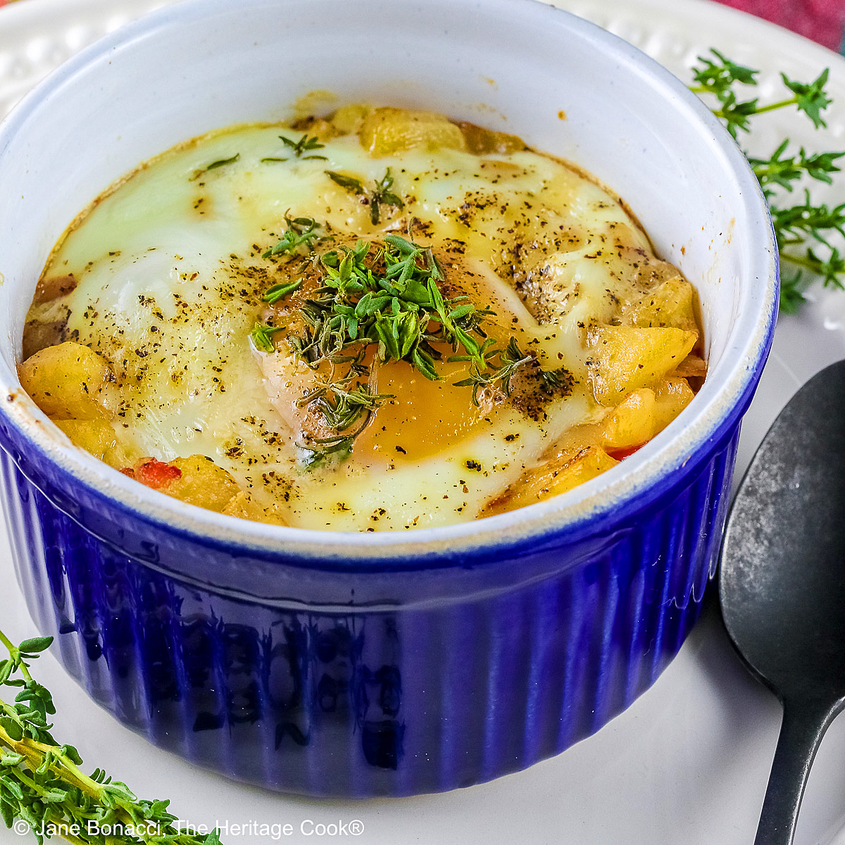 Provençal baked eggs in a potato nest in dark blue ramekins on a baking sheet with a Silpat to stop sliding, sitting on a multi-colored cloth with a black spoon © 2026 Jane Bonacci, The Heritage Cook.