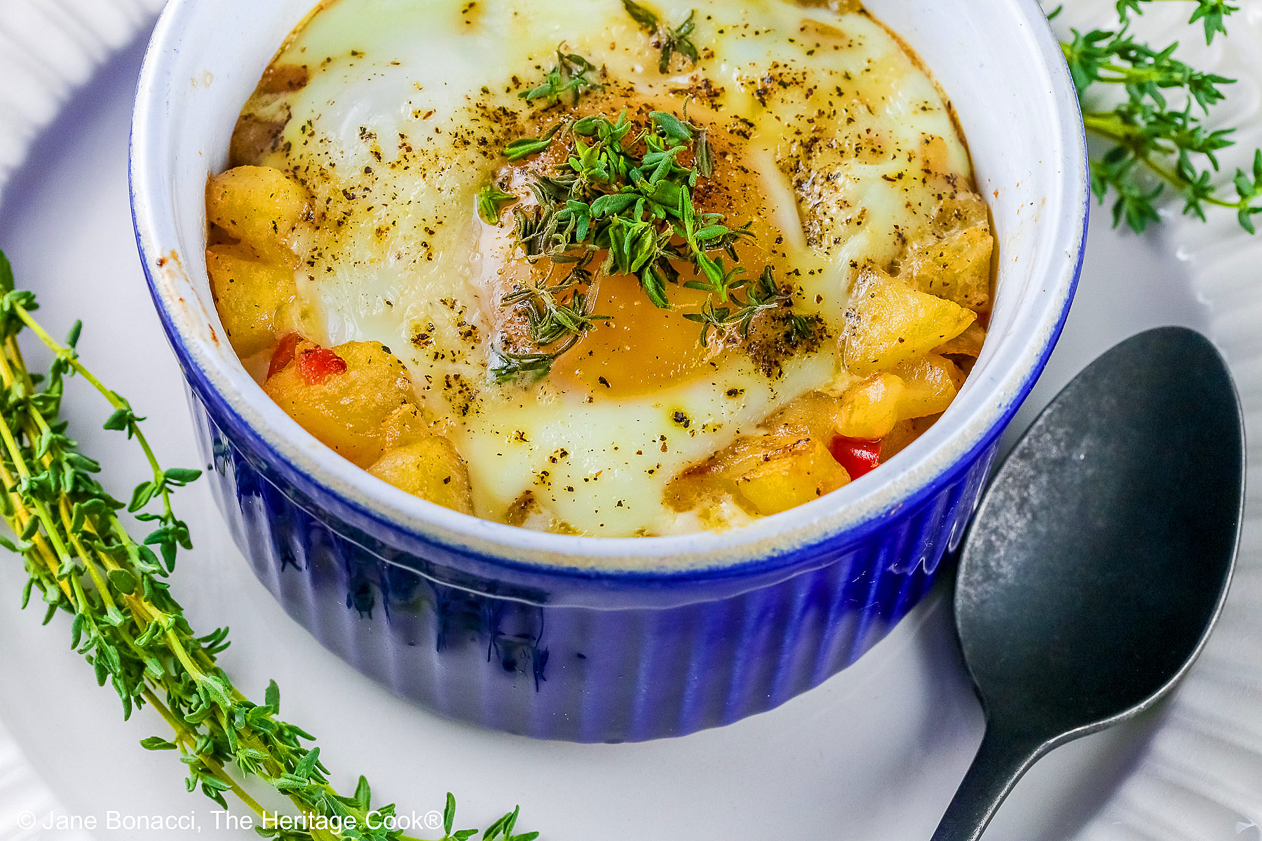 Provençal baked eggs in a potato nest in dark blue ramekins on a baking sheet with a Silpat to stop sliding, sitting on a multi-colored cloth with a black spoon © 2026 Jane Bonacci, The Heritage Cook.