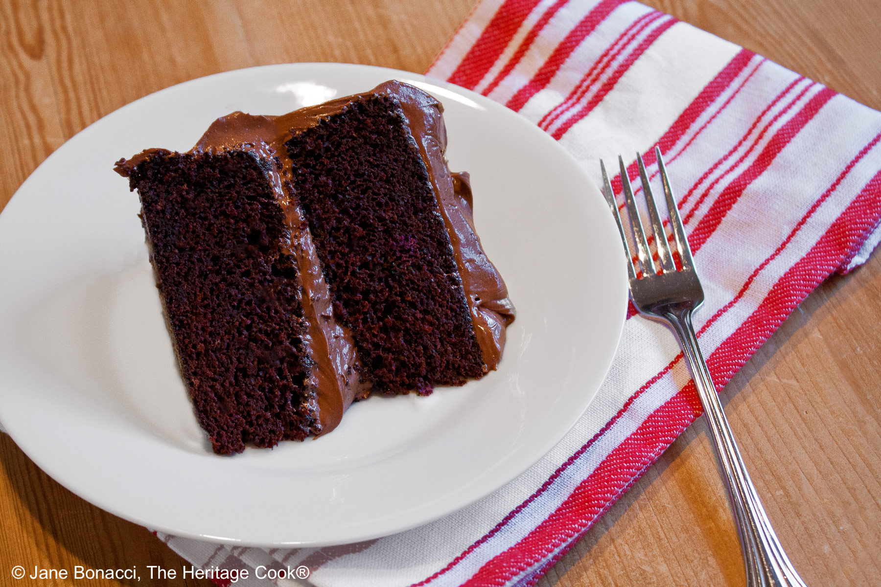 Slice of Dark Chocolate Layer Cake with Chocolate Frosting on a white plate with a red and white striped cloth and a fork © 2024 Jane Bonacci, The Heritage Cook. 