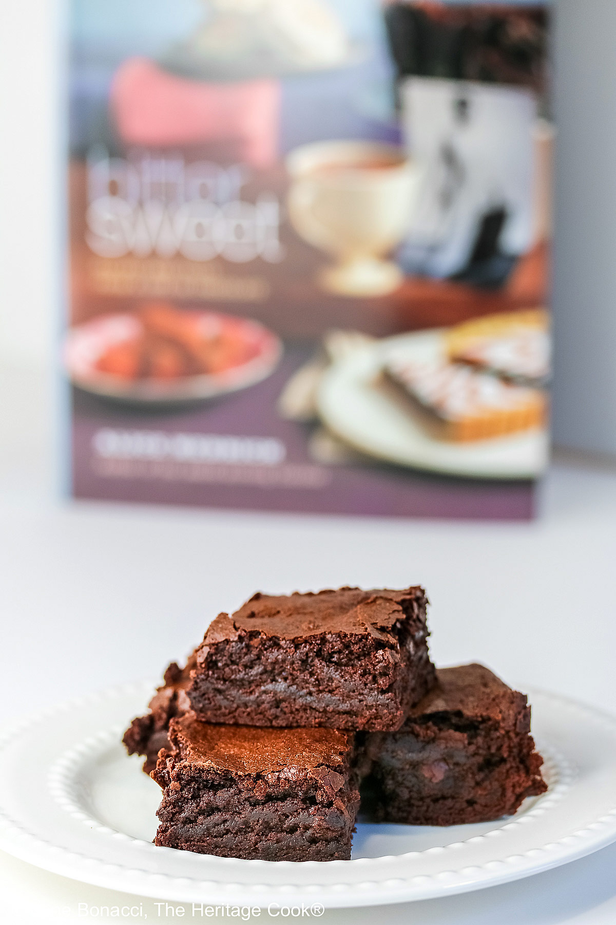 The Best Chewy Brownies, rich and so chocolatey, stacked on a white plate with two rows of white pearls, sitting on a white background; some shots of the cookbook, “Bittersweet” by Alice Medrich; © 2025 Jane Bonacci, The Heritage Cook.