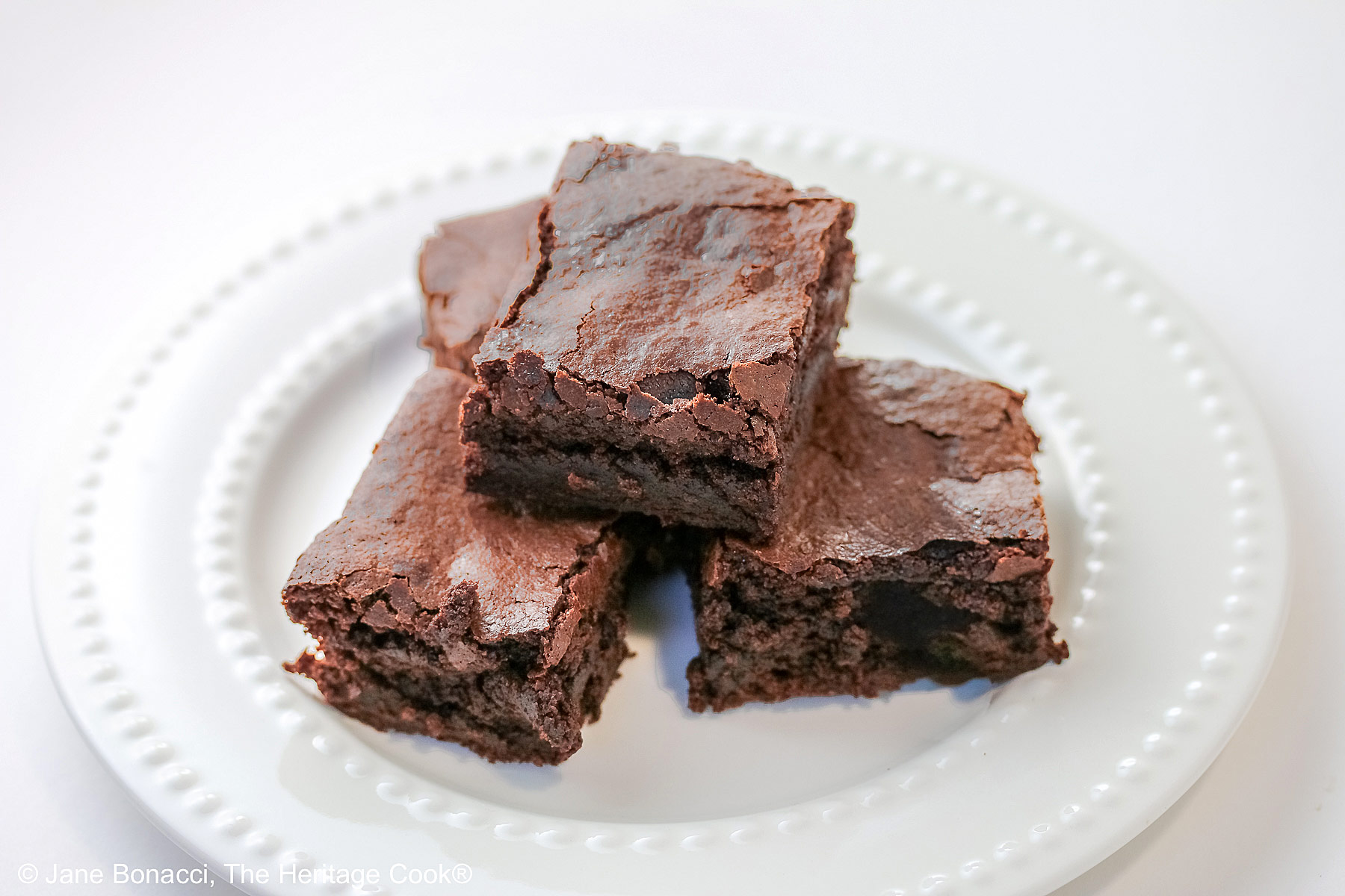 The Best Chewy Brownies, rich and so chocolatey, stacked on a white plate with two rows of white pearls, sitting on a white background; some shots of the cookbook, “Bittersweet” by Alice Medrich; © 2025 Jane Bonacci, The Heritage Cook.