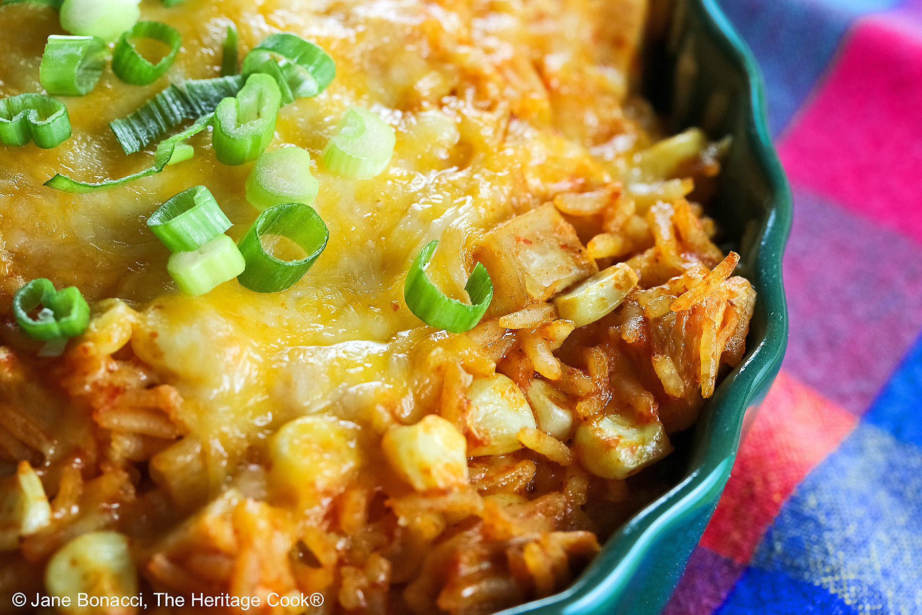Red tinged rice piled in a green au gratin dish with melted cheese over top and sprinkled with sliced green onions, some with a spoon on the side © 2025 Jane Bonacci, The Heritage Cook.