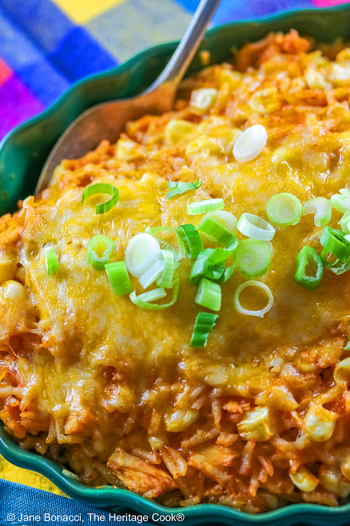 Red tinged rice piled in a green au gratin dish with melted cheese over top and sprinkled with sliced green onions, some with a spoon on the side © 2025 Jane Bonacci, The Heritage Cook.
