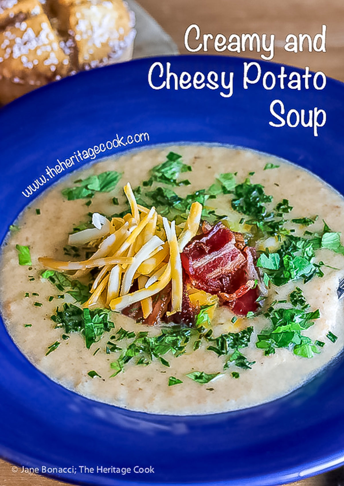 Deep blue bowls brimming with hot Creamy Cheesy Potato Soup topped with bacon, shredded cheese, green chiles, parsley and chopped green onions on a wooden table with a blue and white cloth alongside © 2025 Jane Bonacci, The Heritage Cook.