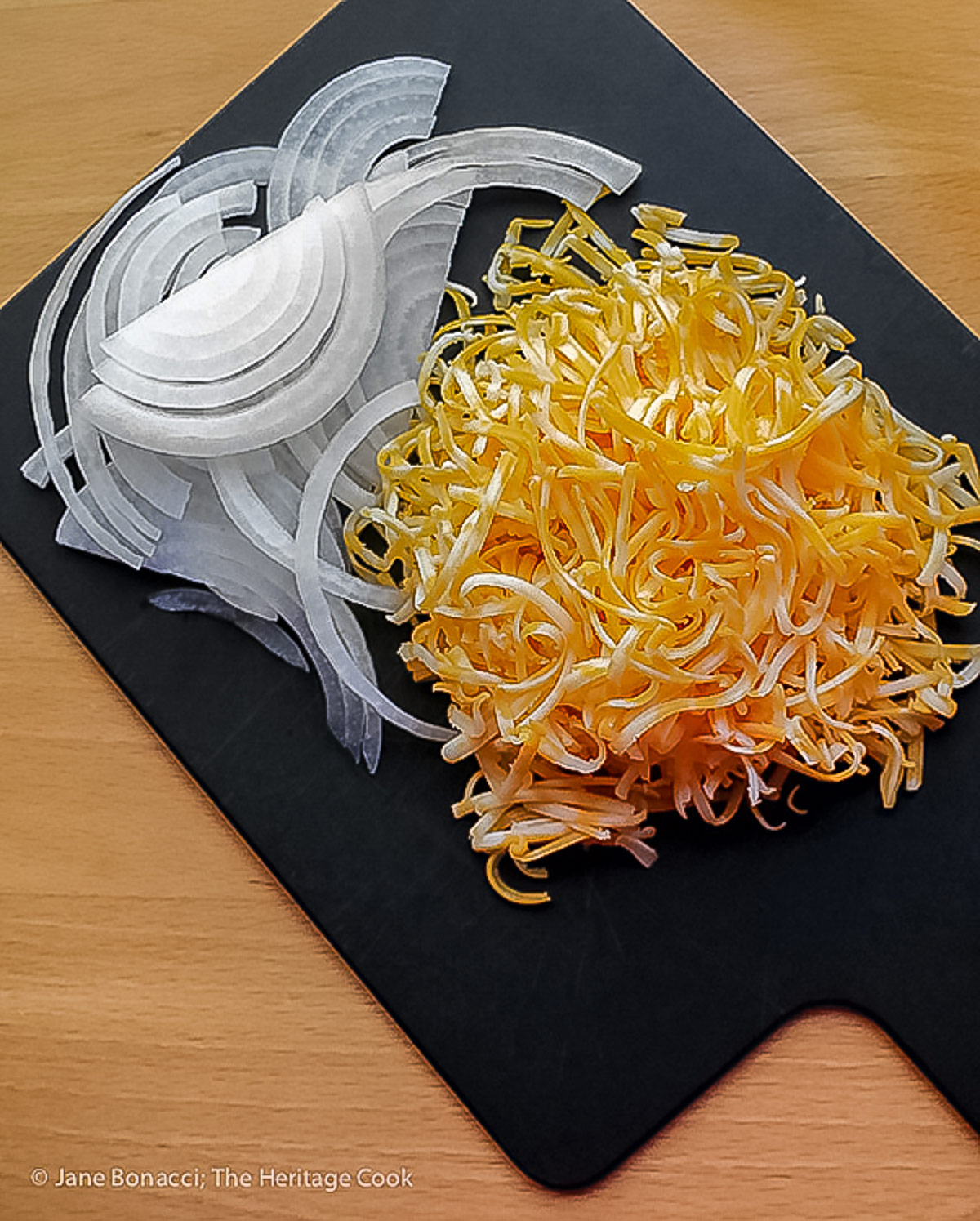 Sliced onions and grated cheddar cheese on a small black cutting board. 