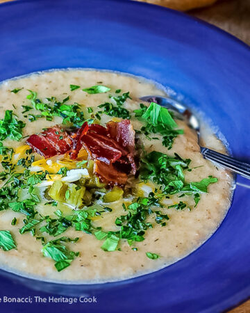 Deep blue bowls brimming with hot Creamy Cheesy Potato Soup topped with bacon, shredded cheese, green chiles, parsley and chopped green onions on a wooden table with a blue and white cloth alongside © 2025 Jane Bonacci, The Heritage Cook.