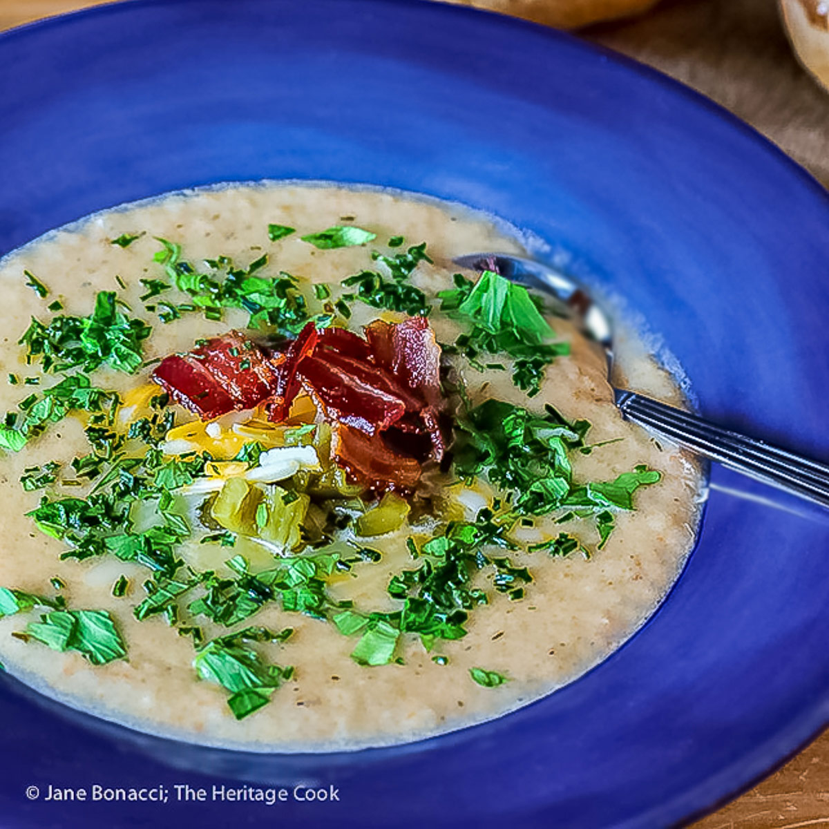 Deep blue bowls brimming with hot Creamy Cheesy Potato Soup topped with bacon, shredded cheese, green chiles, parsley and chopped green onions on a wooden table with a blue and white cloth alongside © 2025 Jane Bonacci, The Heritage Cook. 