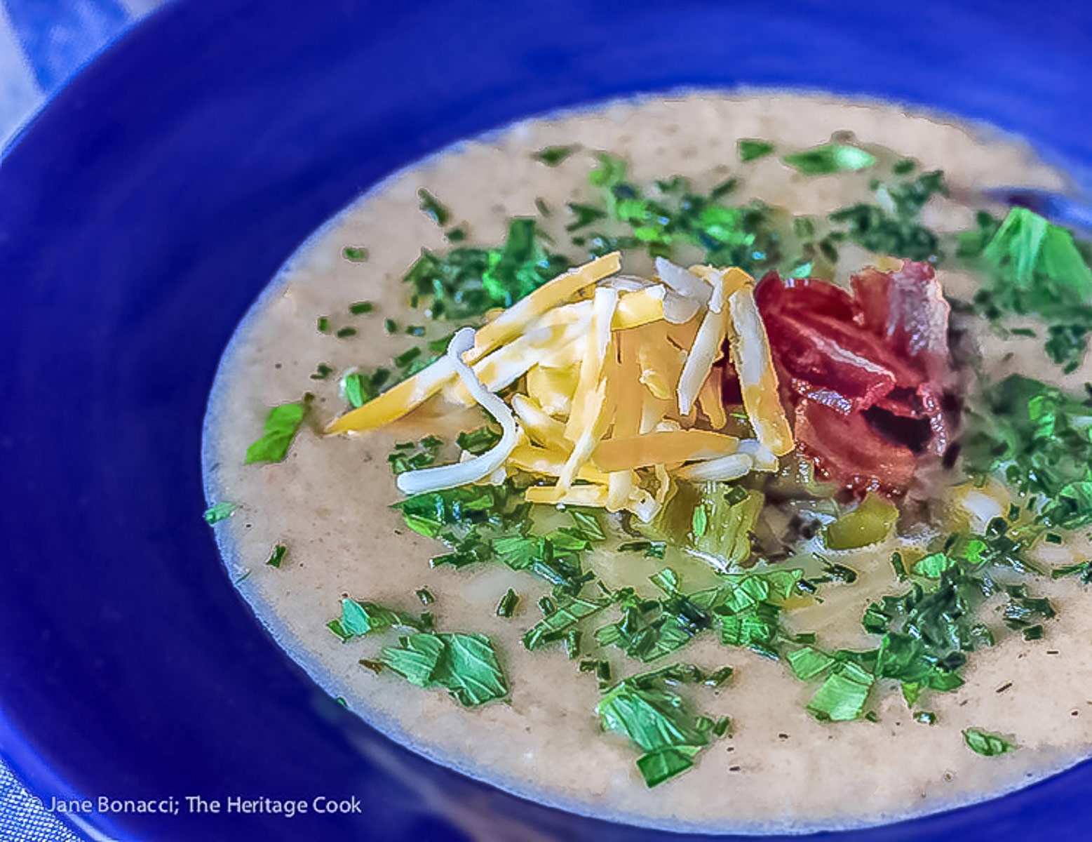 Deep blue bowls brimming with hot Creamy Cheesy Potato Soup topped with bacon, shredded cheese, green chiles, parsley and chopped green onions on a wooden table with a blue and white cloth alongside © 2025 Jane Bonacci, The Heritage Cook. 