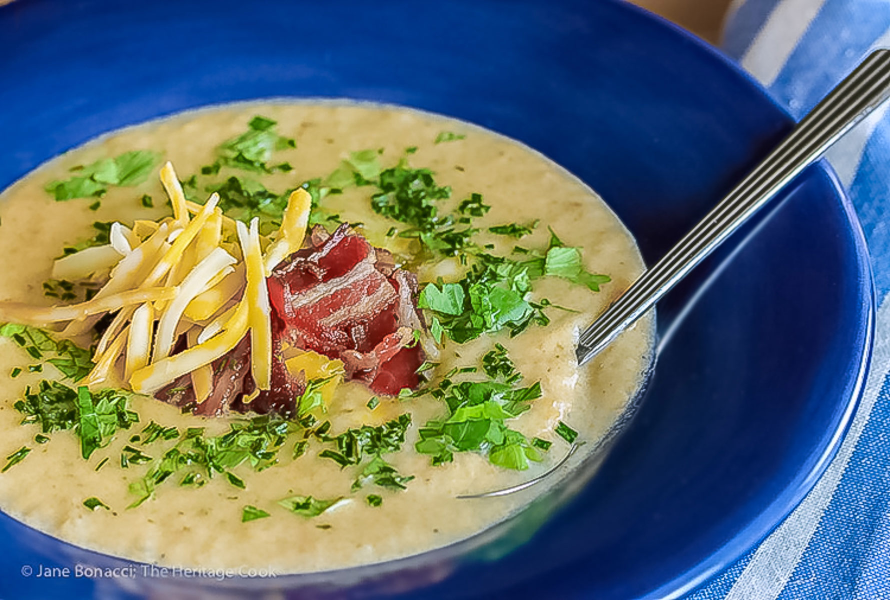 Deep blue bowls brimming with hot Creamy Cheesy Potato Soup topped with bacon, shredded cheese, green chiles, parsley and chopped green onions on a wooden table with a blue and white cloth alongside © 2025 Jane Bonacci, The Heritage Cook. 