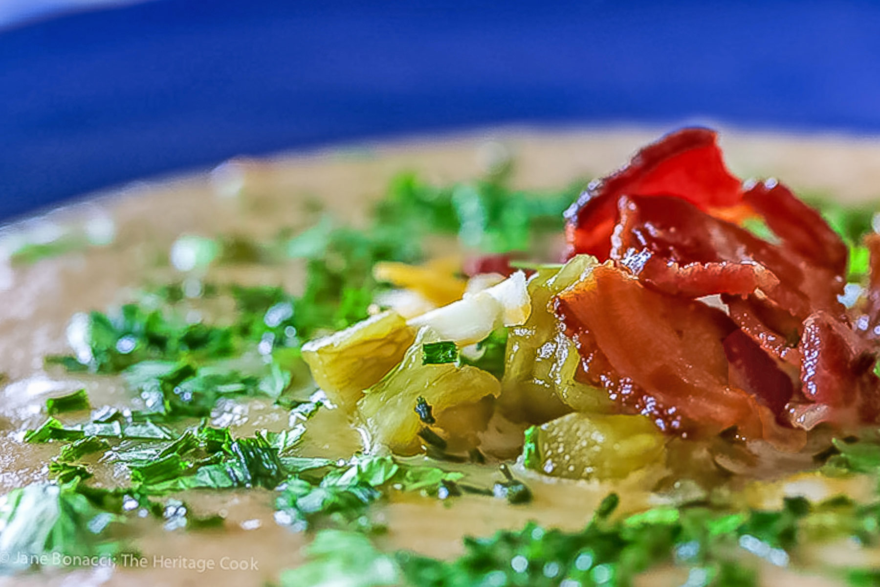 Deep blue bowls brimming with hot Creamy Cheesy Potato Soup topped with bacon, shredded cheese, green chiles, parsley and chopped green onions on a wooden table with a blue and white cloth alongside © 2025 Jane Bonacci, The Heritage Cook. 