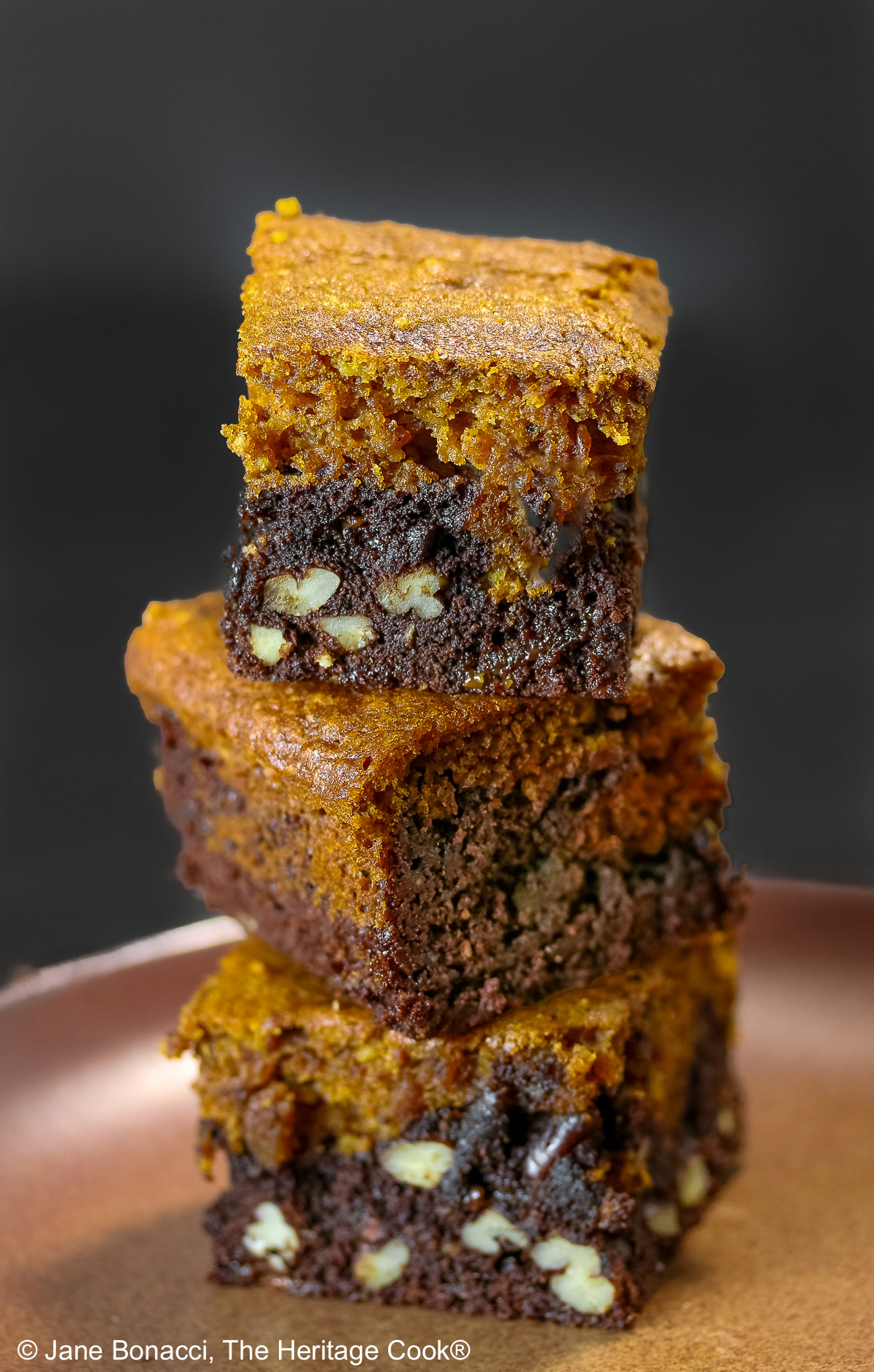 Pumpkin Swirl Brownies cut into squares and sitting on a dark plate, some stacked 3 high with a dark background that highlights the contrast between the dark brownies on the bottom with a topping of golden pumpkin cake on each piece © 2026 Jane Bonacci, The Heritage Cook.