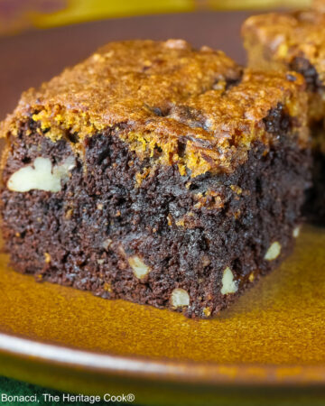Pumpkin Swirl Brownies cut into squares and sitting on a dark plate, some stacked 3 high with a dark background that highlights the contrast between the dark brownies on the bottom with a topping of golden pumpkin cake on each piece © 2026 Jane Bonacci, The Heritage Cook.
