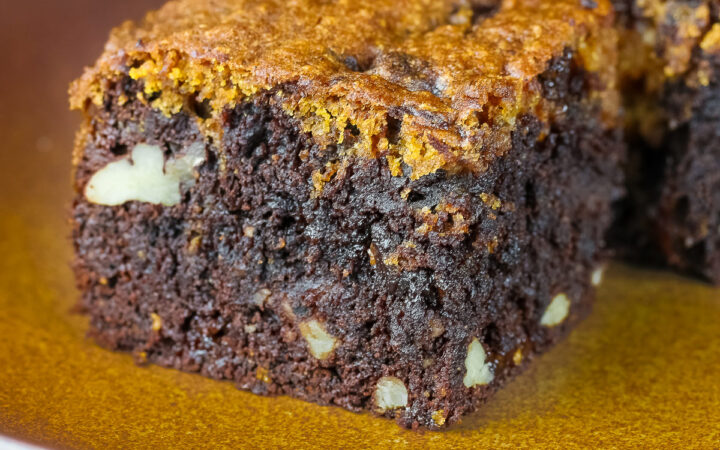 Pumpkin Swirl Brownies cut into squares and sitting on a dark plate, some stacked 3 high with a dark background that highlights the contrast between the dark brownies on the bottom with a topping of golden pumpkin cake on each piece © 2026 Jane Bonacci, The Heritage Cook.