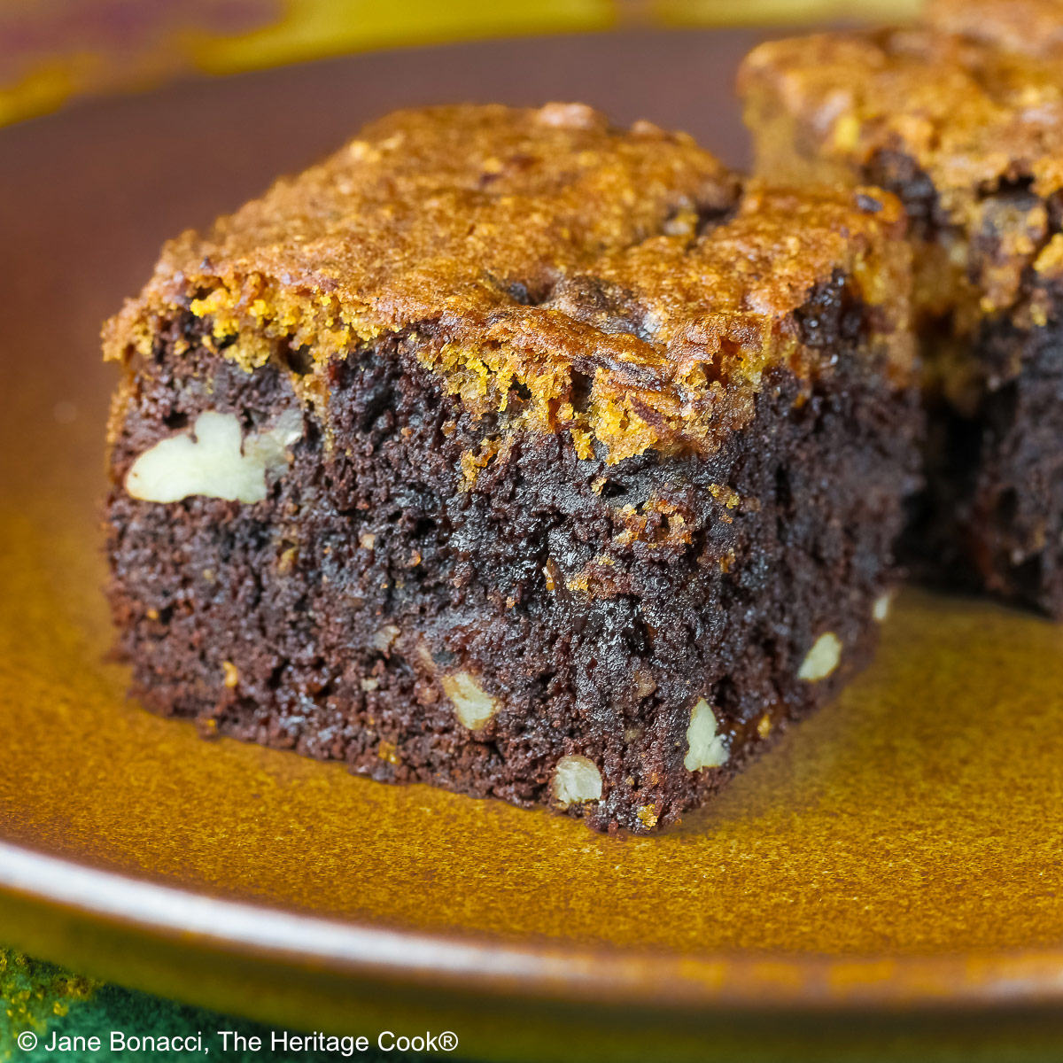 Pumpkin Swirl Brownies cut into squares and sitting on a dark plate, some stacked 3 high with a dark background that highlights the contrast between the dark brownies on the bottom with a topping of golden pumpkin cake on each piece © 2026 Jane Bonacci, The Heritage Cook.